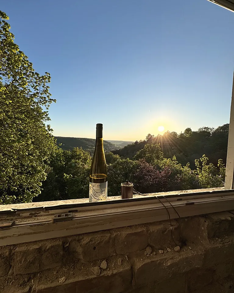 Eine Flasche Wein auf der Fensterbank im Waldhaus, dahinter die Sonne über dem Tal