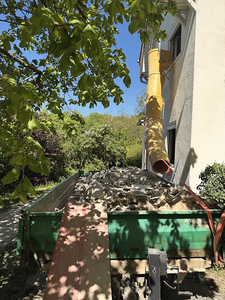 Container mit gelber Bauschuttrutsche an der Fassade des Waldhauses, daneben blühender Flieder